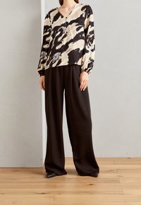 Sort og beige mønstret bluse med lange ærmer og knapdetaljer, parret med brede sorte bukser i et glat stof, på et lyst trægulv.