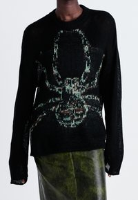 Zwarte gebreide trui met een groot groen en bruin spinnenafbeelding, met een losse pasvorm en een gestructureerd patroon. Gemaakt in combinatie met een groene leren rok.