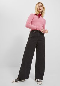 Maglietta rosa a maniche lunghe, jeans neri a gamba larga e sneakers nere con suola bianca. Dettagli notevoli includono polsini a coste e orlo sfilacciato.