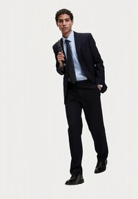 Mörk skräddarsydd kostym med ljusblå skjorta och mönstrad slips. Personen håller en svart väska och bär blanka svarta skor.