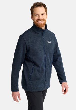 Lächelnder Mann mit roten Haaren, der eine dunkelblaue Jack Wolfskin Fleecejacke mit Reißverschluss und schwarze Hose trägt, vor einem einfarbigen Hintergrund posiert.