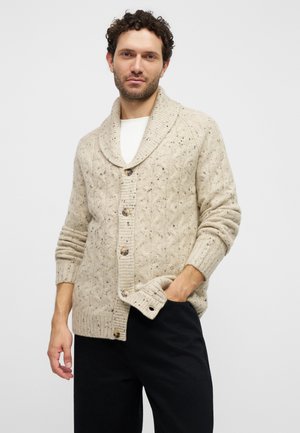 Un homme porte un cardigan crème moucheté à torsades sur une chemise blanche et un pantalon noir. Il se tient debout, une main dans la poche.