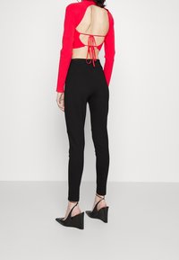 Vrouw in een rode lange mouw top met open rug, vastgebonden met touwtjes, zwarte hoge taille broek en zwarte wedge hakken, staand tegen een effen achtergrond.