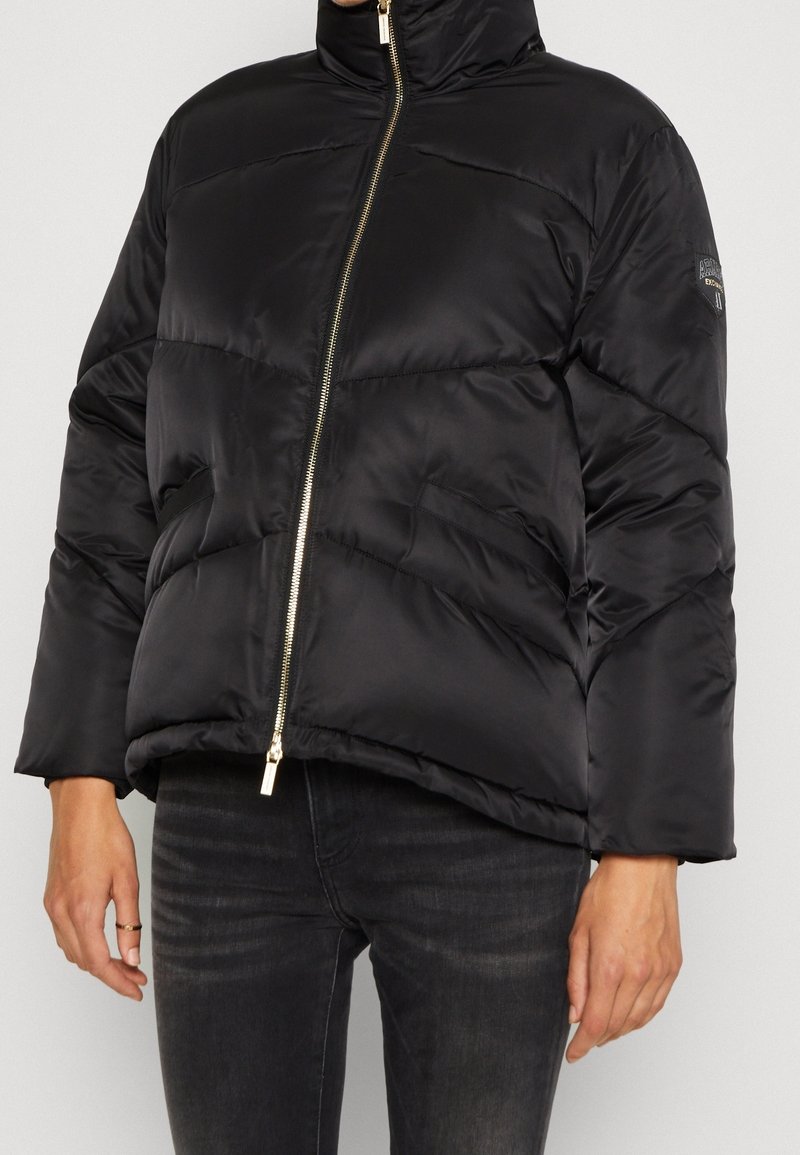 Veste matelassée noire avec fermeture éclair dorée, col montant, poches latérales, portée sur un jean foncé, main gauche visible avec une bague au doigt du milieu.