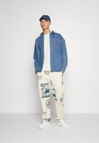 Τζην πουκάμισο, λευκό μπλουζάκι, κρεμ cargo παντελόνι με μπλε γραφικά μοτίβα και λευκά αθλητικά παπούτσια. Τα αξεσουάρ περιλαμβάνουν ένα μπλε καπέλο.