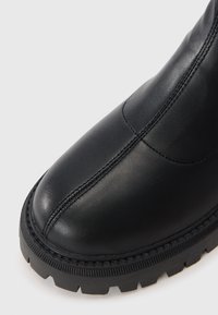 Zwarte leren enkelboot met een ronde neus, zichtbare stiksels en een dikke, gestructureerde rubberen zool met een profielpatroon.