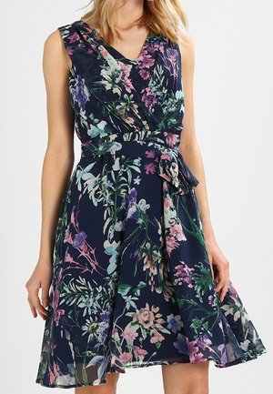 Ärmelloses marineblaues Kleid mit pinkem, lila und grünem Blumenmuster, V-Ausschnitt und gebundenem Taillengürtel, getragen von einer stehenden Person.