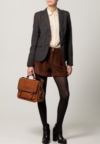 Femme portant un blazer gris, une chemise beige à boutons, un short en velours côtelé marron, des collants noirs, des bottines noires, tenant un sac à main en cuir marron avec un motif floral.