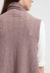 Strickweste ohne Ärmel in einem sanften Mauve-Ton, mit hohem Kragen, gerippter Textur und offenem Rückendesign, kombiniert mit einem weißen Langarmshirt.