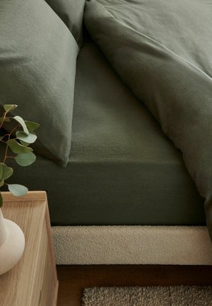 Nahaufnahme eines ungemachten Bettes mit dunkelgrünen Laken und Kissen neben einem hölzernen Nachttisch mit einer Vase mit grünen Blättern.