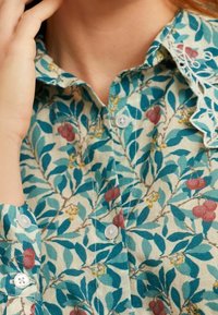 Chemise à boutons florale avec un fond crème clair, ornée de feuilles vertes et de baies rouges, en tissu texturé et avec un col festonné.