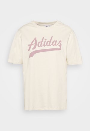 Kremfarvet T-shirt med korte ærmer og lyserød, kursiv "Adidas"-logo med understregning tværs over brystet, vist mod en ensfarvet baggrund.