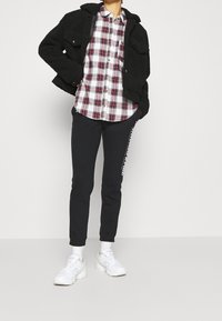 Giacca nera e vaporosa indossata sopra una camicia a quadri bianca e rossa, abbinata a pantaloni sportivi neri con testo bianco e sneakers bianche.
