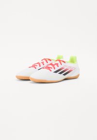 Scarpe da calcio bianche con superficie testurizzata, vivaci lacci rossi, accentuazioni gialle e suola in gomma, con tre strisce nere di lato.