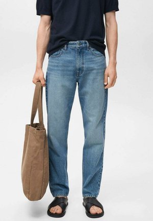 Jeans in denim azzurro chiaro con vestibilità rilassata, abbinati a una t-shirt blu navy e sandali neri. Una borsa tote in tessuto marrone è tenuta in una mano.