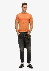 Camiseta gráfica naranja con texto, combinada con jeans grises. Camisa de cuadros atada a la cintura, usando mocasines negros y calcetines blancos.