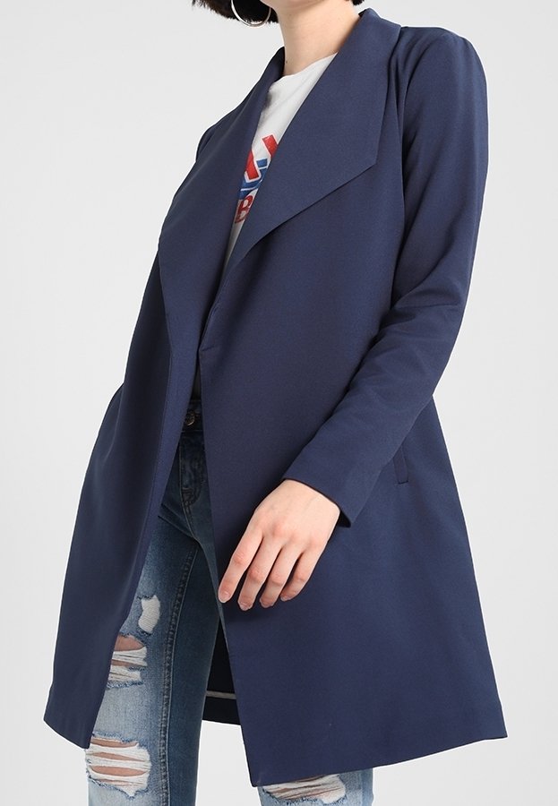 Personne portant un manteau long bleu marine sur un t-shirt graphique blanc et un jean déchiré bleu clair, se tenant devant un fond uni.