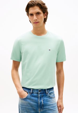 Leicht mintgrünes Baumwoll-T-Shirt mit kurzen Ärmeln und Rundhalsausschnitt. Mit einem kleinen, auf der Brust gestickten Logo. Getragen zu ausgewaschenen blauen Jeans.