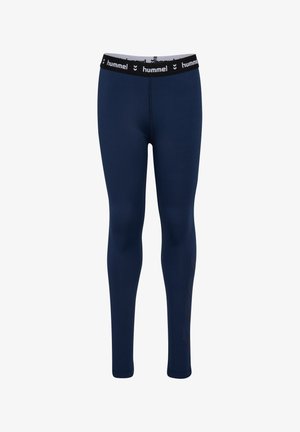 Ung pige i navyblåt sportstøj med armene krydset, stående med hvide sokker og navy sneakers mod en ensfarvet baggrund.