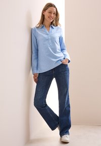 Camicia a righe azzurro chiaro con scollo a V e maniche lunghe, abbinata a jeans a zampa d'elefante blu scuro e sneakers bianche.