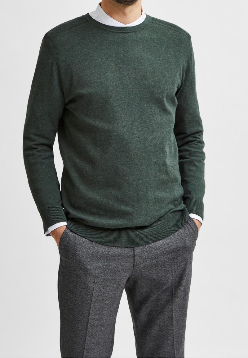 Maglione verde scuro con scollo rotondo, polsini e orlo a costine, indossato sopra una camicia bianca con colletto, abbinato a pantaloni grigi con una texture.