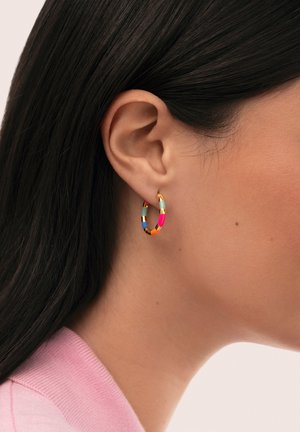 Mujer que lleva un pequeño aro de oro con segmentos de esmalte multicolor, mostrado en primer plano en su oreja y cuello izquierdo.