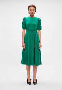 Πράσινο φόρεμα μέχρι το γόνατο με κοντά, φουσκωτά μανίκια, σφιγμένη μέση δεμένη με αντίστοιχο ζωνάκι, απαλό υλικό και με μετριότητα στον λαιμό. Μαύρες γόβες.