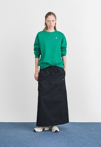 Grønn sweatshirt med ribbestrikkede mansjetter, paret med en svart maxiskjørt med cargolommer. Hvite sneakers fullfører antrekket.