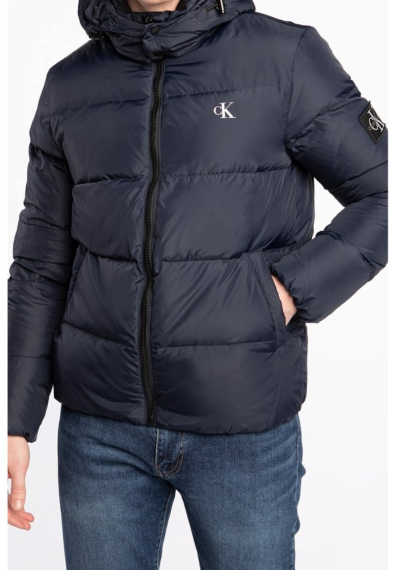 Námořnická modrá bunda s kapucí a logem cK na hrudi a rukávu, nošená s modrými džíny, ruka v pravé kapse, přední zip zapnutý.