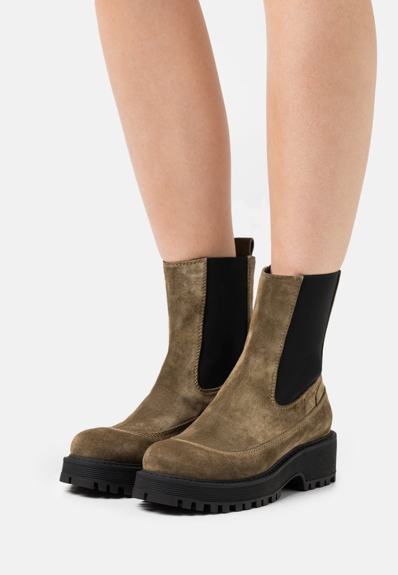 Zamszowe botki za kostkę w oliwkowej zieleni z czarnymi elastycznymi panelami bocznymi i grubymi czarnymi podeszwami z protektorem, noszone na gołych nogach.