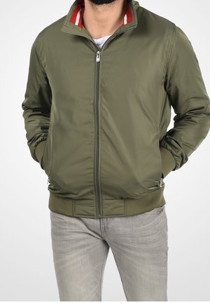 Mand iført en olivengrøn lynlåstrøje med ribstrikkede manchetter og linning, kombineret med lysegrå jeans, hænderne i lommerne.