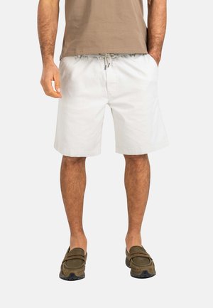 Hombre con camiseta beige, pantalones cortos blancos hasta la rodilla con cordón y zapatos marrones sin cordones, de pie con las manos en los bolsillos.