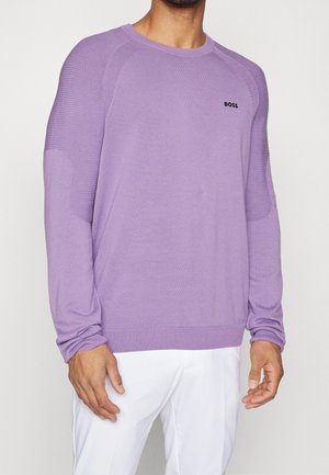 Mężczyzna w lawendowym swetrze z długim rękawem, z prążkowanymi detalami na ramionach i czarnym logo "BOSS", w połączeniu z białymi spodniami na jednolitym tle.