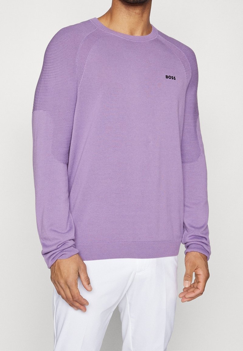 Homme portant un pull lavender à manches longues avec des détails côtelés aux épaules et le logo noir "BOSS", associé à un pantalon blanc sur un fond uni.