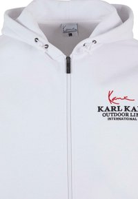 Felpa bianca con zip realizzata in tessuto morbido, dotata di cappuccio con coulisse e logo ricamato in rosso e nero sul lato sinistro del petto.