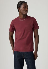 Bordeauxrotes Baumwoll-T-Shirt mit kurzen Ärmeln und einer Brusttasche. Verfügt über ein kleines Logo-Label auf der Tasche. Kombiniert mit dunkelblauen Jeans.