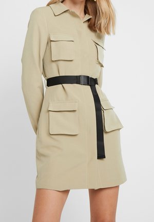 Beżowa sukienka koszulowa z długim rękawem, czterema kieszeniami z patkami i czarnym paskiem podkreślającym talię, noszona przez stojącą osobę o blond włosach.