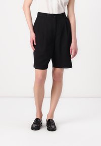 Schwarze, maßgeschneiderte Shorts aus glattem Stoff mit hoher Taille, ausgestattet mit einem Knopfverschluss vorne und seitlichen Taschen. Kombiniert mit schwarzen Slippern.