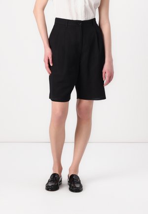 Pantaloni shorts neri su misura realizzati in tessuto morbido con vita alta, dotati di chiusura frontale a bottone e tasche laterali. Abbinati a mocassini neri.