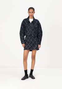 Jeansjack met geometrisch stikselpatroon, gecombineerd met een bijpassende rok. Donkerblauwe kleur, cropped pasvorm en klassieke knoopsluiting. Zwarte loafers.