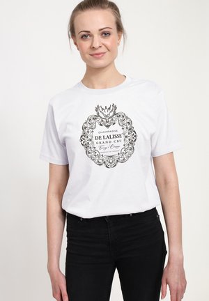 Jonge vrouw met licht haar in een staart draagt een wit T-shirt met een sierlijk zwart tekstontwerp "Champagne De Lalisse Grand Cru", gecombineerd met zwarte spijkerbroek.