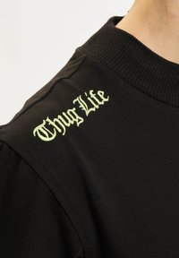Černá krátkozápasová košile vyrobená z bavlny, s zeleně vyšitým textem "Thug Life" na rameni blízko límce.