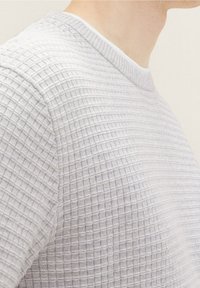 Maglietta a maglia testurizzata grigia chiara con scollo rotondo, indossata da una persona mostrata dal collo alla parte superiore del torso.