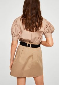 Top beige con maniche a sbuffo e vita aderente, abbinato a una gonna beige plissettata. La gonna presenta una cintura nera. Sfondo semplice.