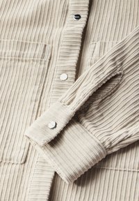 Chemise en velours côtelé beige avec une texture côtelée verticale, dotée de boutons-pression argentés et de deux poches poitrine. Tissu doux et confortable.