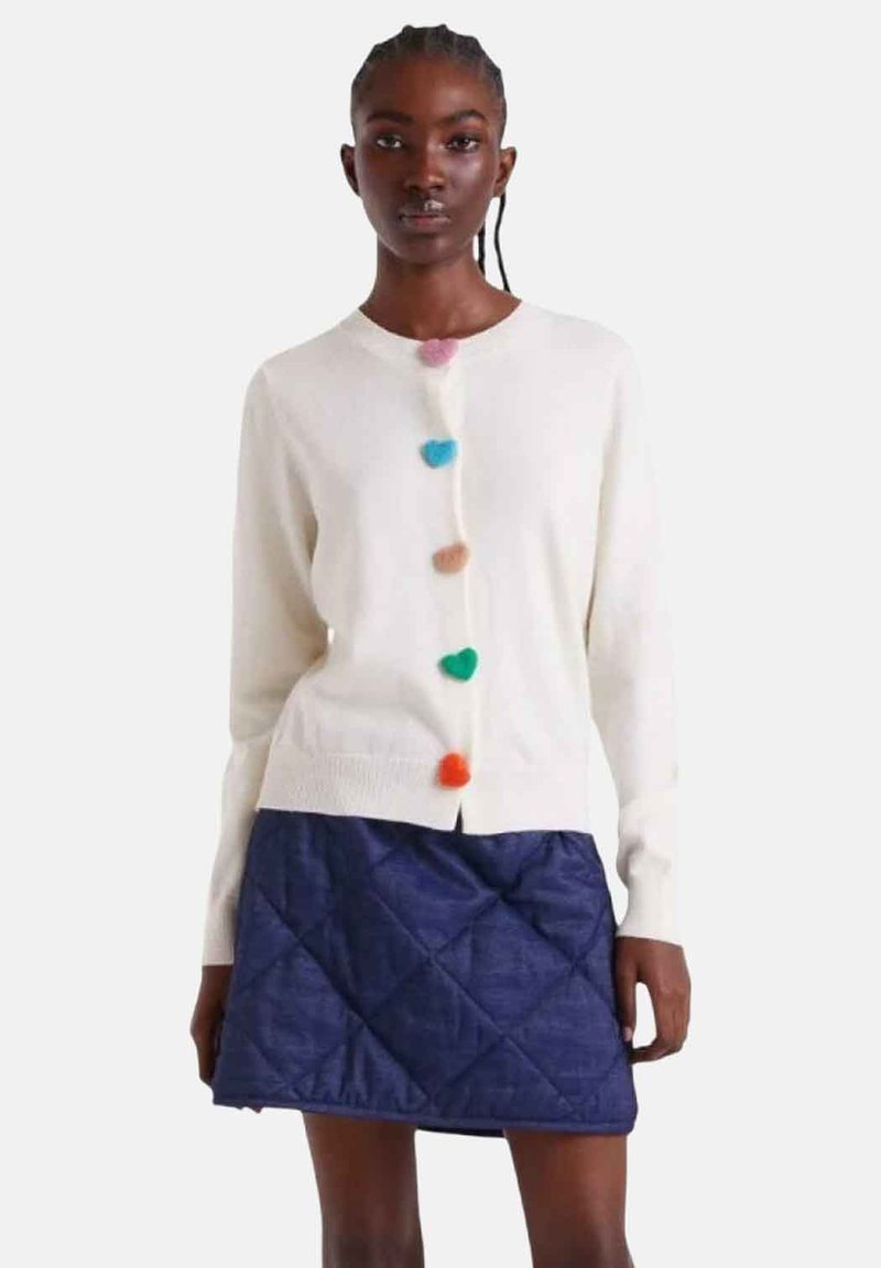 Weißer Cardigan mit bunten Pompon-Knöpfen und V-Ausschnitt, kombiniert mit einem blauen, gesteppte Minirock mit Diamantmuster.