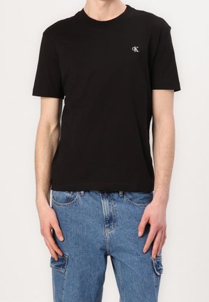 Mężczyzna w jednolitym czarnym t-shircie z krótkim rękawem, na którym po lewej stronie piersi znajduje się małe białe logo "cK", oraz w niebieskich dżinsowych spodenka na cargo.
