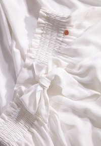 Witte stof met een gladde textuur heeft geplooide details aan de ene kant, accentueert door een strik en een klein oranje label.