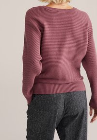 Rukávový svetr s dlouhými rukávy v barvě šeříku s kulatým výstřihem a texturovaným povrchem. Kombinován se šedými kalhotami s jemným tečkovaným vzorem.