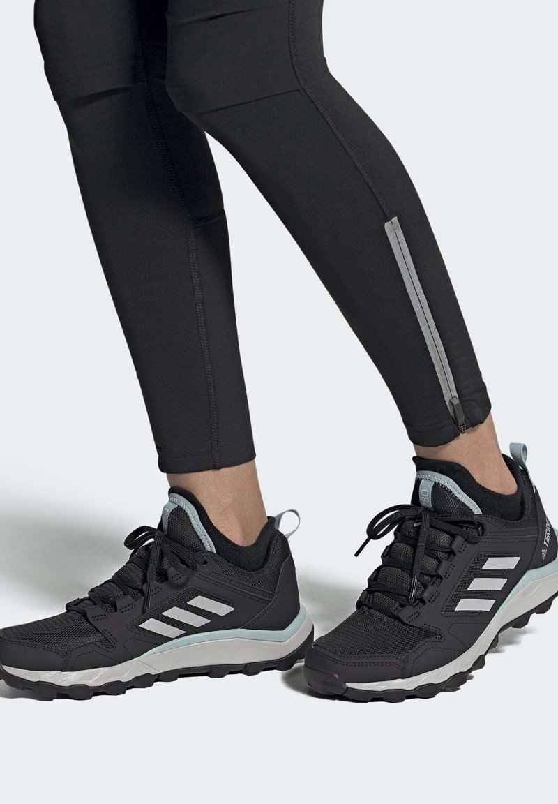 Jambes portant des leggings de sport noirs avec des fermetures éclair aux chevilles et des chaussures de trail noires avec des bandes blanches et des accents bleu clair.